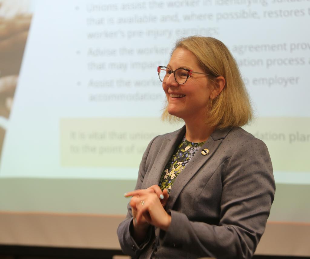 Tracey Newman presenting at a podium in front of a screen with text on it