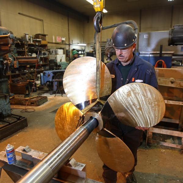 A person working in a workshop, holding a large metal propeller with four blades attached to a long shaft. 