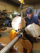 A person working in a workshop, holding a large metal propeller with four blades attached to a long shaft. 