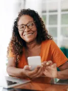 A person with curly hair wearing an orange shirt, sitting at a table and holding a smartphone while using a laptop.
