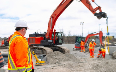 Photograph of a construction site featuring workers in bright orange safety gear and white helmets operating heavy machinery, including two large red excavators.