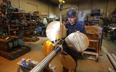 A person working in a workshop, holding a large metal propeller with four blades attached to a long shaft. 